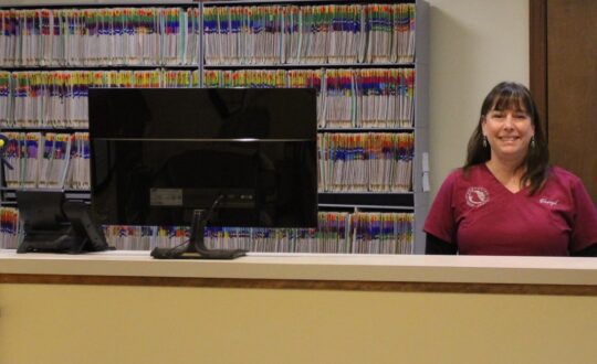 A smiling white woman with shoulder length dark hair and bangs in a maroon Berkshire Humane Society scrub top stands behind a yellow counter with a large computer screen on it. Shelves of records are behind her.