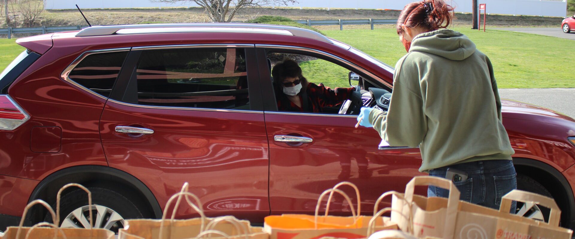 A person takes an order from a maroon SUV with bags of food in foreground