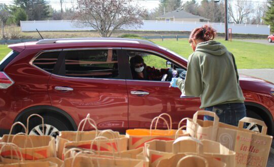 A person takes an order from a maroon SUV with bags of food in foreground
